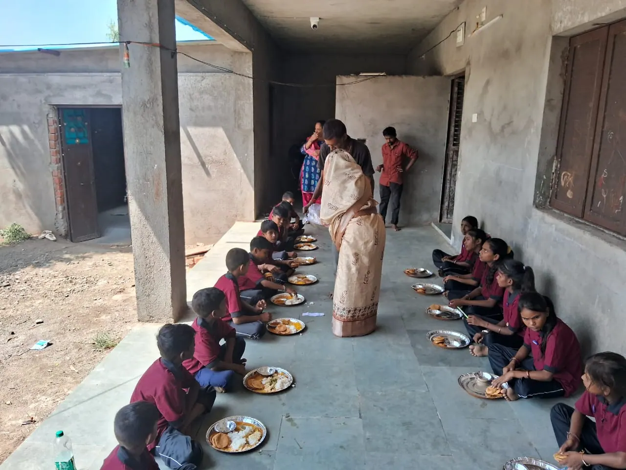 A staff member serving food to students seated on the verandah floor at mealtime.